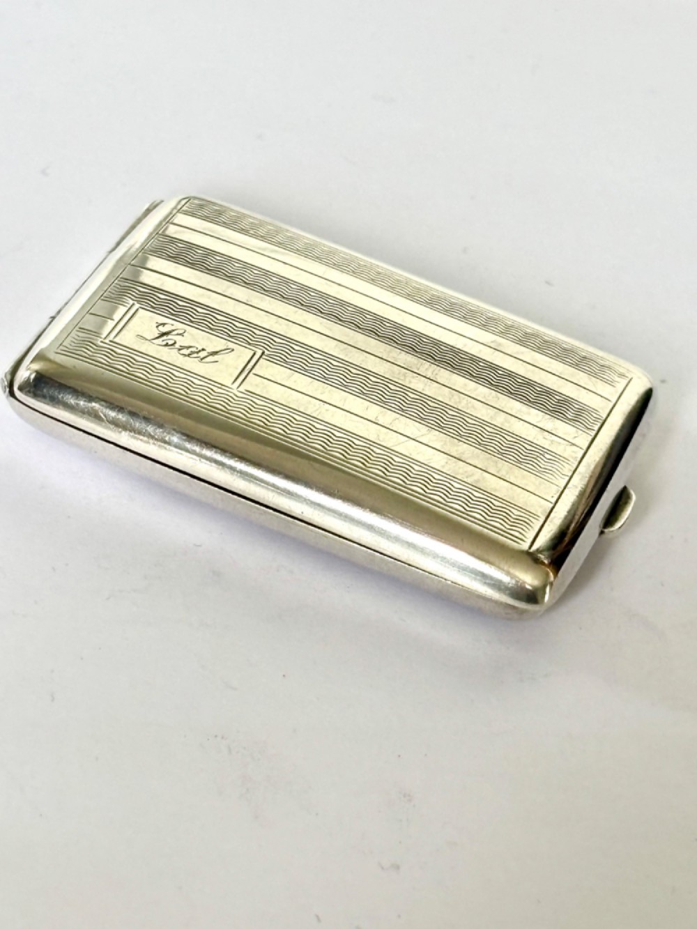 Rectangular sterling silver matchbook case featuring engine-turned geometric linear pattern and a central monogrammed cartouche.