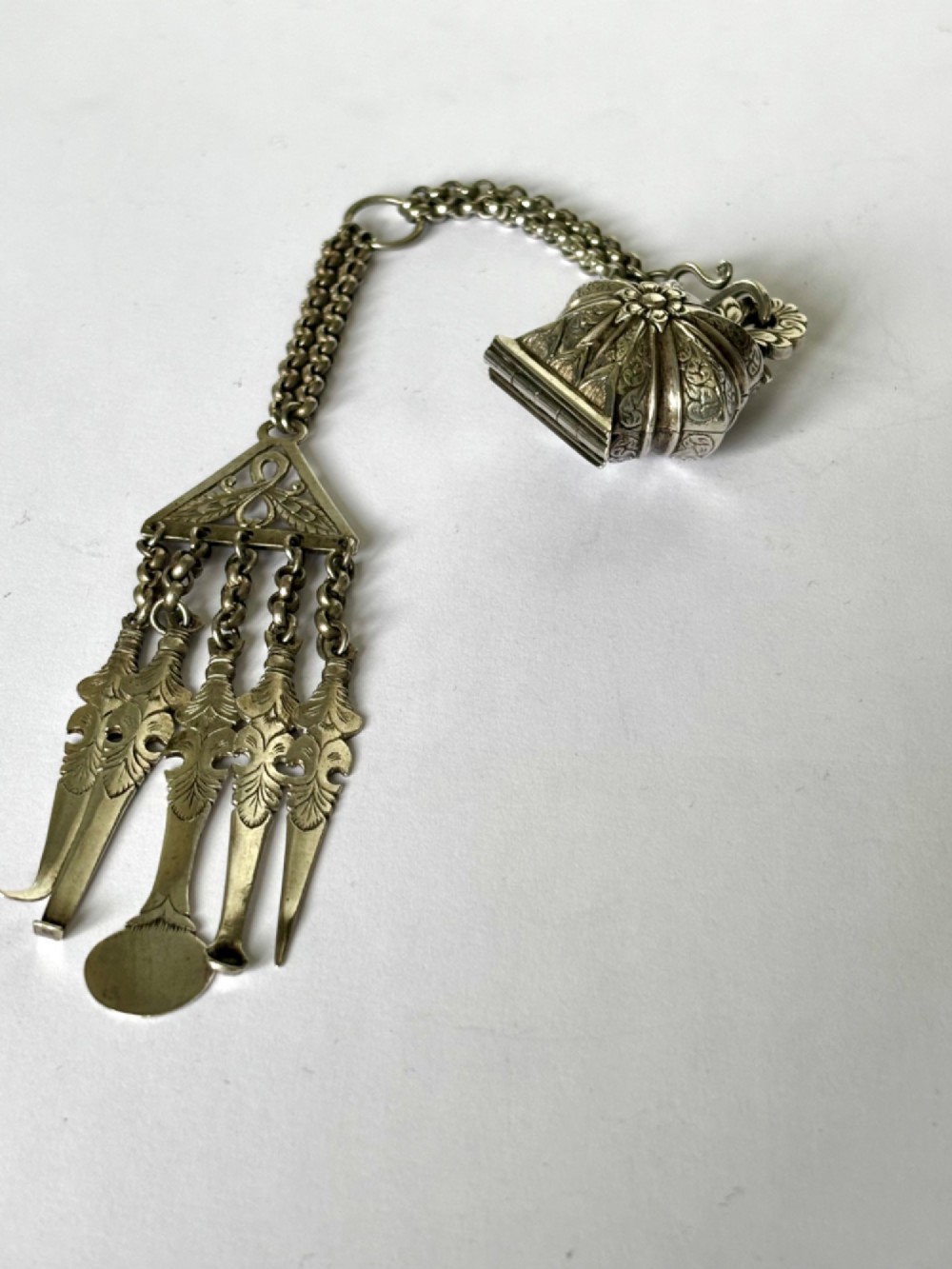 An antique silver white metal chatelaine featuring an ornate clip connected by links to five suspended grooming tools and a fluted vinaigrette container.