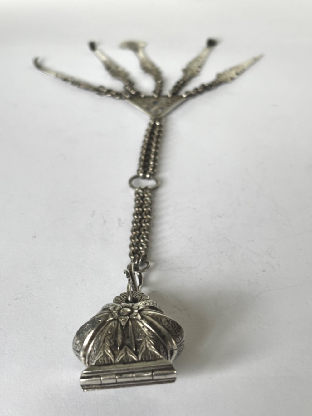 An antique silver white metal chatelaine featuring an ornate clip connected by links to five suspended grooming tools and a fluted vinaigrette container. View II