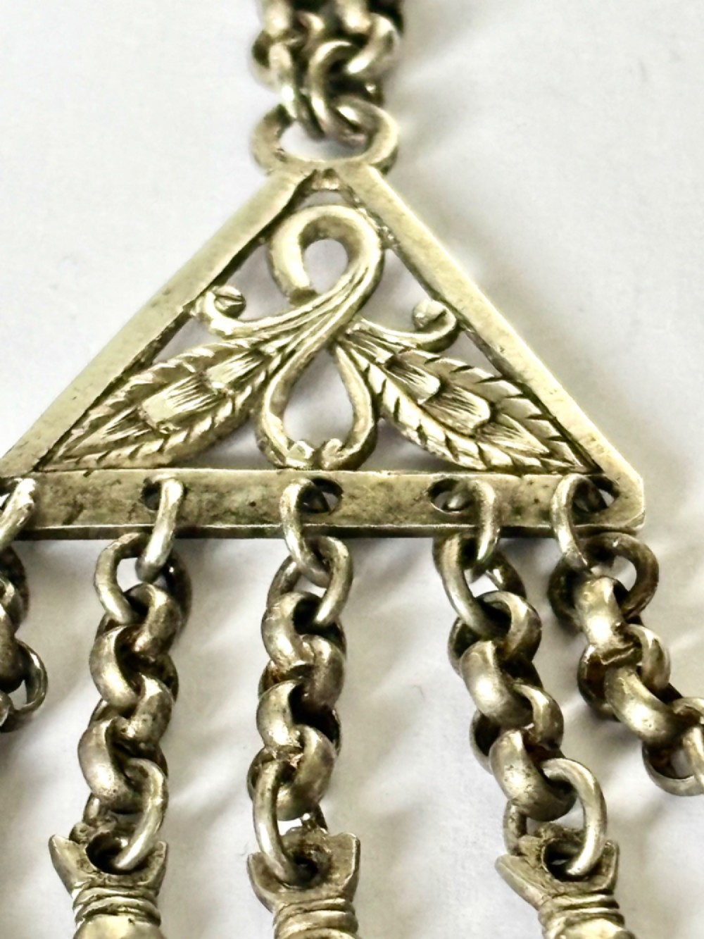 An antique silver white metal chatelaine featuring an ornate clip connected by links to five suspended grooming tools and a fluted vinaigrette container. View X