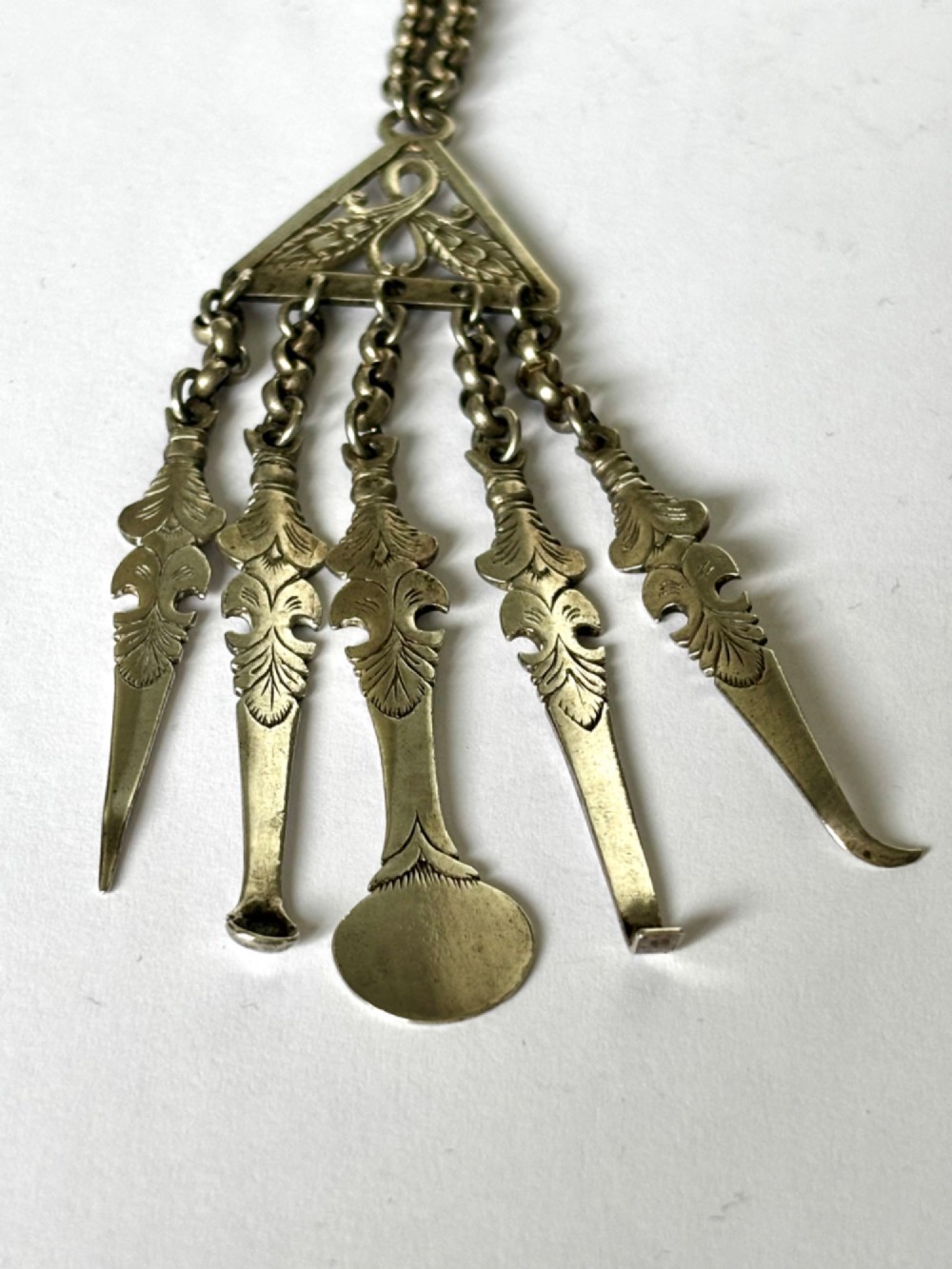 An antique silver white metal chatelaine featuring an ornate clip connected by links to five suspended grooming tools and a fluted vinaigrette container. View XI