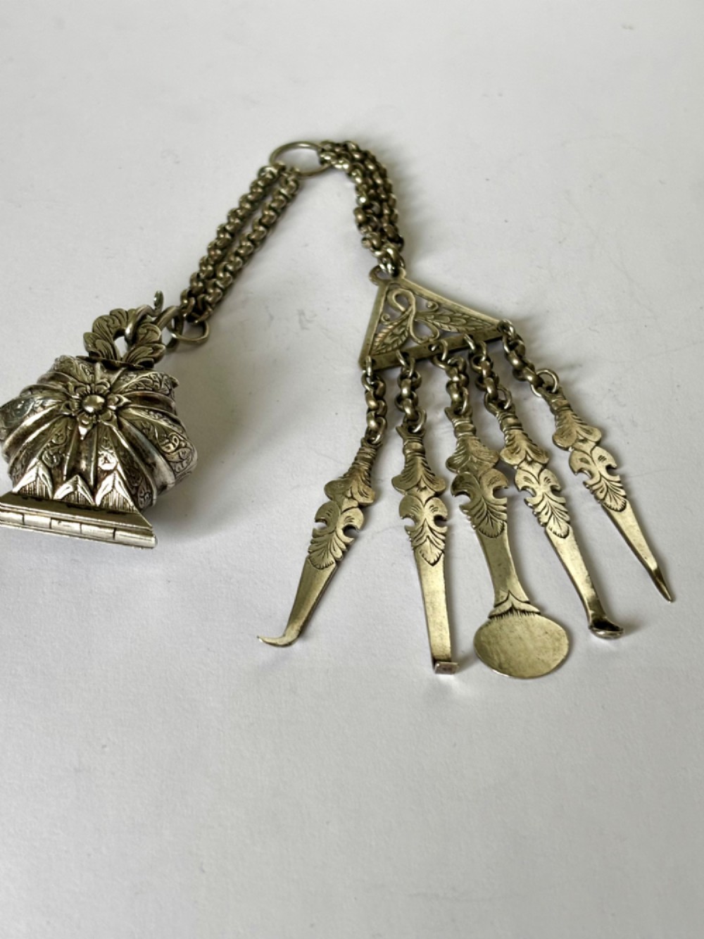 An antique silver white metal chatelaine featuring an ornate clip connected by links to five suspended grooming tools and a fluted vinaigrette container. View XIII