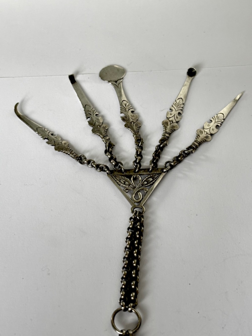 An antique silver white metal chatelaine featuring an ornate clip connected by links to five suspended grooming tools and a fluted vinaigrette container. View XV