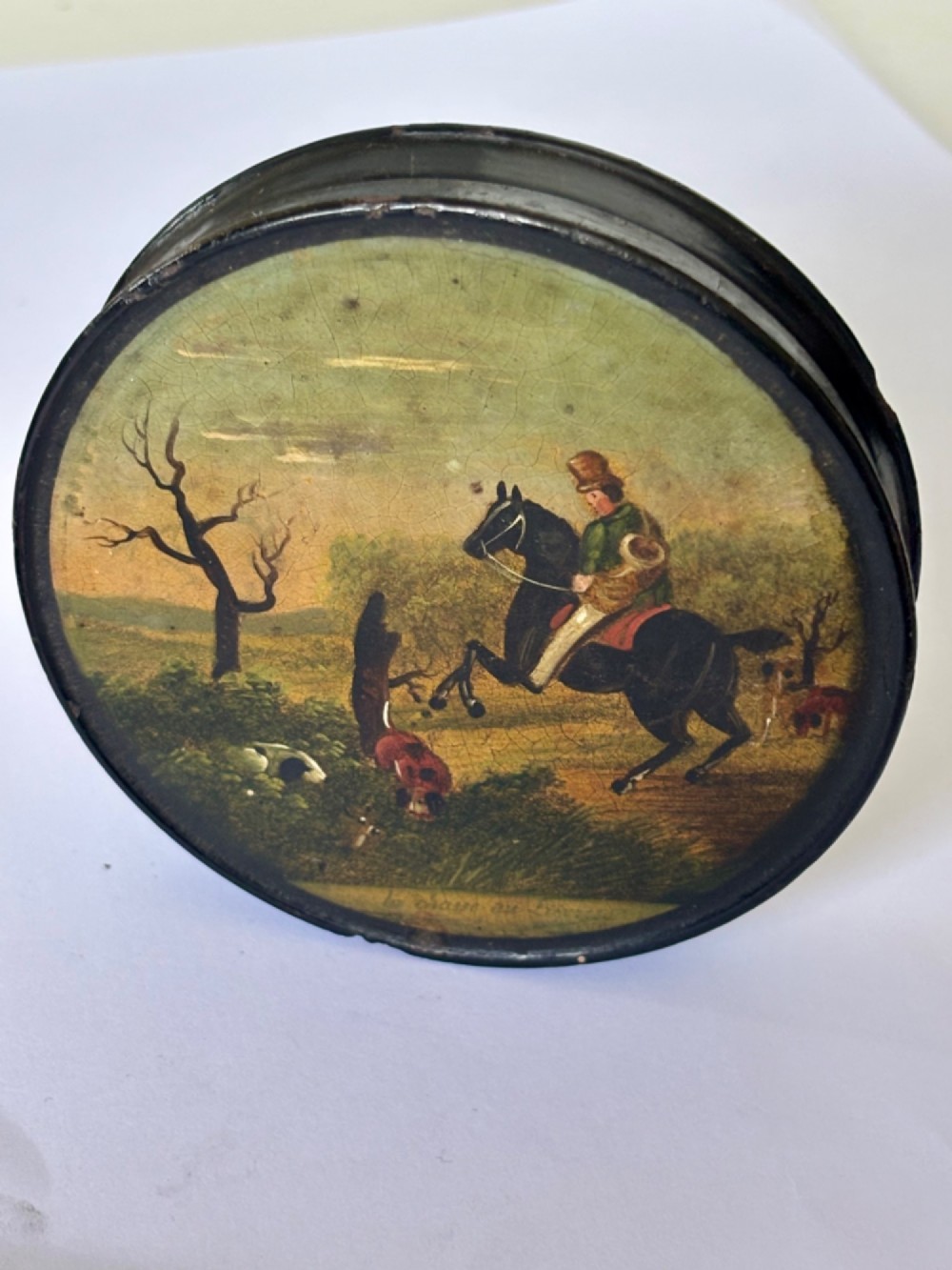 A circular black papier-mache snuff box featuring a hand-painted scene on the lid depicting a horseman and hunting dogs.