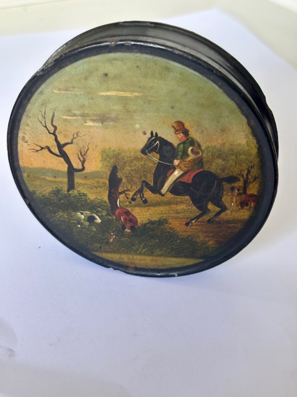 A circular black papier-mache snuff box featuring a hand-painted scene on the lid depicting a horseman and hunting dogs. View II