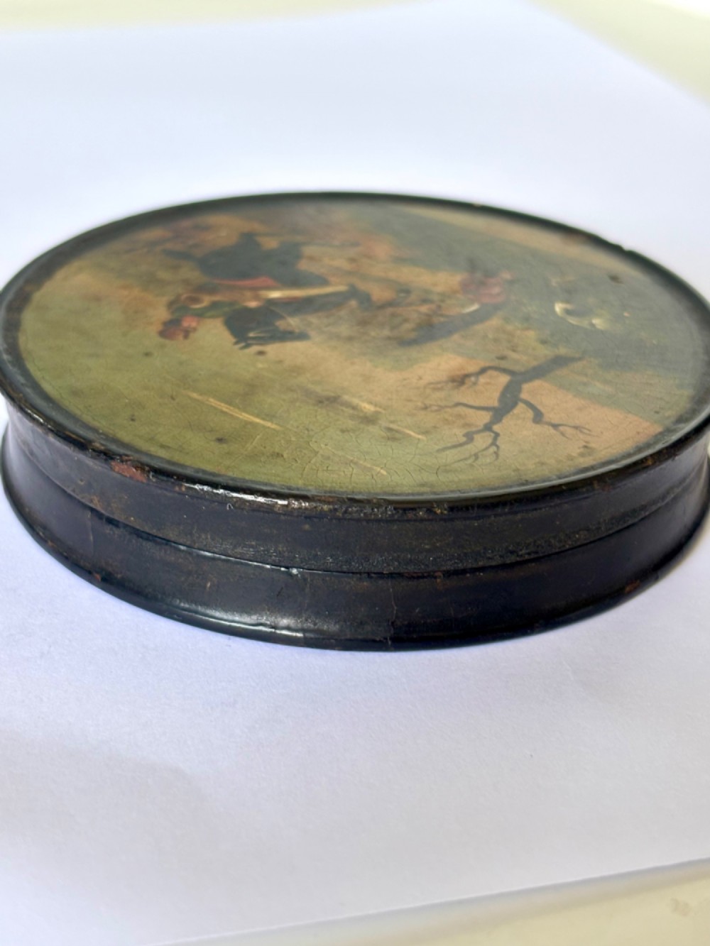 A circular black papier-mache snuff box featuring a hand-painted scene on the lid depicting a horseman and hunting dogs. View VII