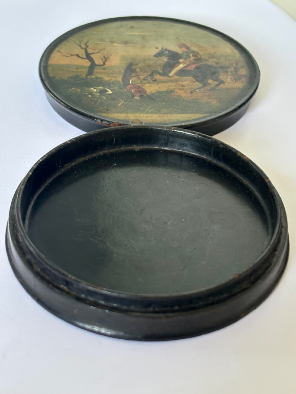 A circular black papier-mache snuff box featuring a hand-painted scene on the lid depicting a horseman and hunting dogs. View VIII