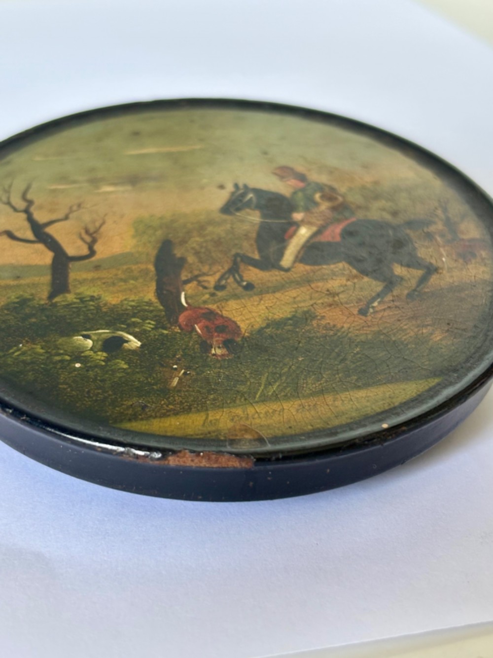 A circular black papier-mache snuff box featuring a hand-painted scene on the lid depicting a horseman and hunting dogs. View IX