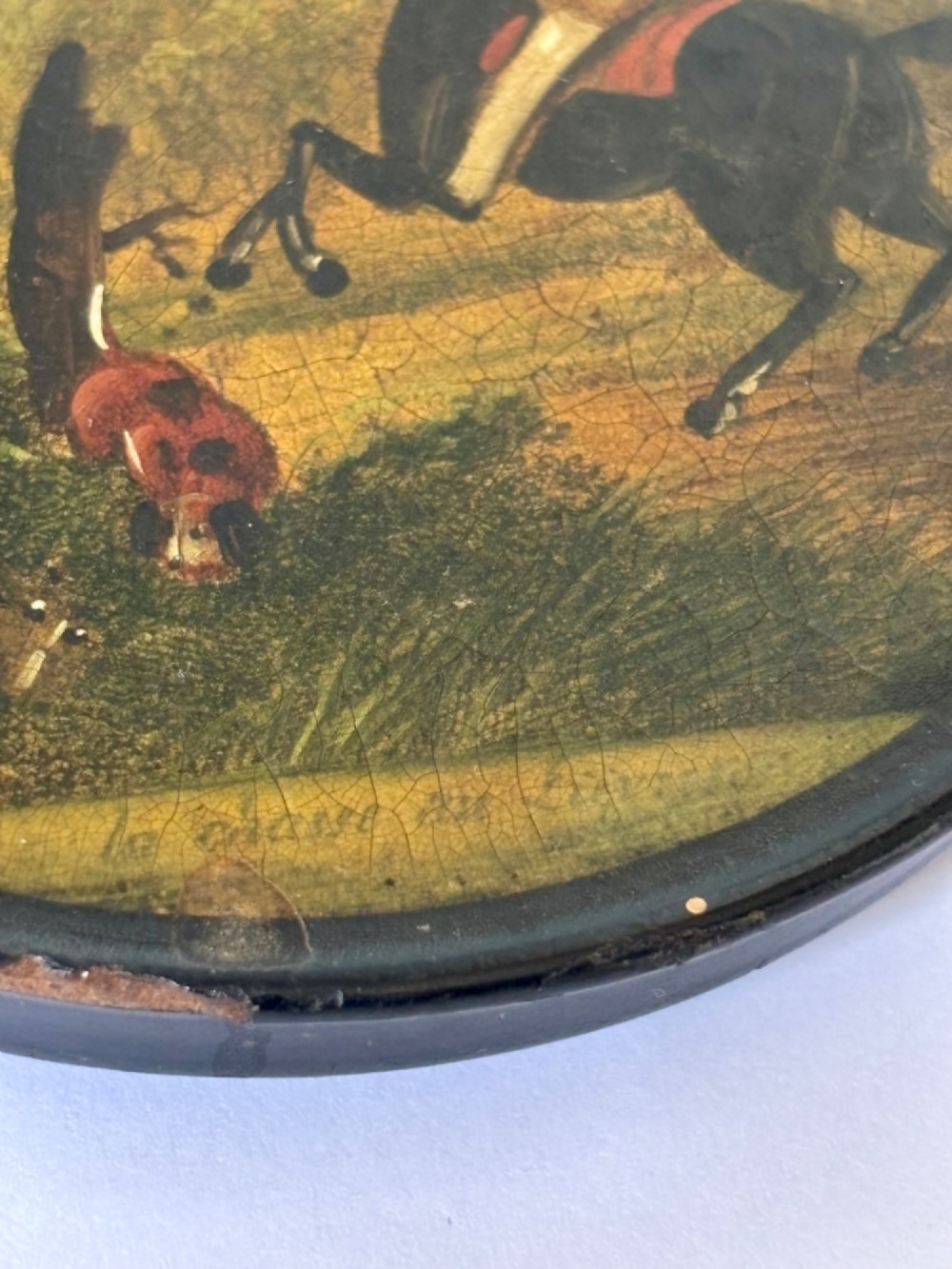 A circular black papier-mache snuff box featuring a hand-painted scene on the lid depicting a horseman and hunting dogs. View X