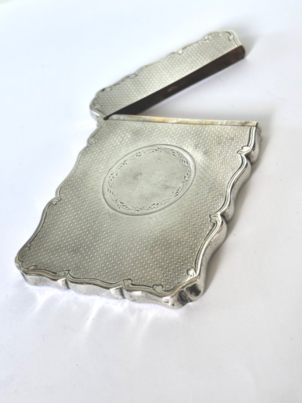 An antique Victorian sterling silver calling card case featuring shaped wavy edges, an engine-turned pattern, and an oval central cartouche.