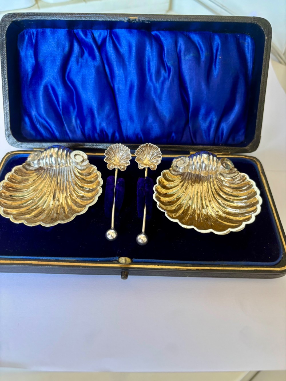 An open fitted presentation case containing two shell-shaped silver salt cellars with gilded interiors and two matching shell-bowl spoons.