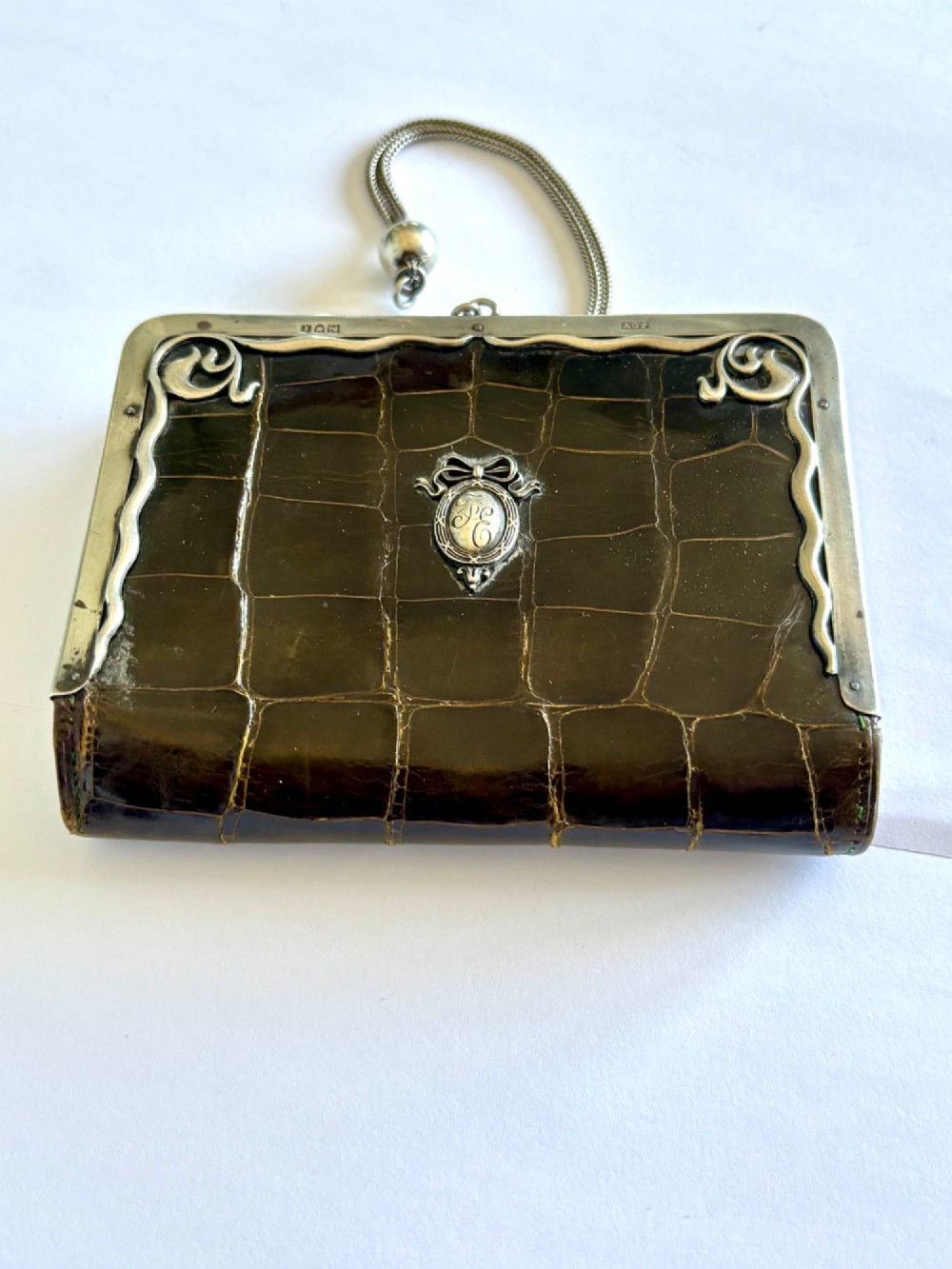 An Art Nouveau dark brown crocodile skin purse featuring hallmarked sterling silver whiplash corner mounts, a central silver cartouche with bow detailing, and a silver chain strap.