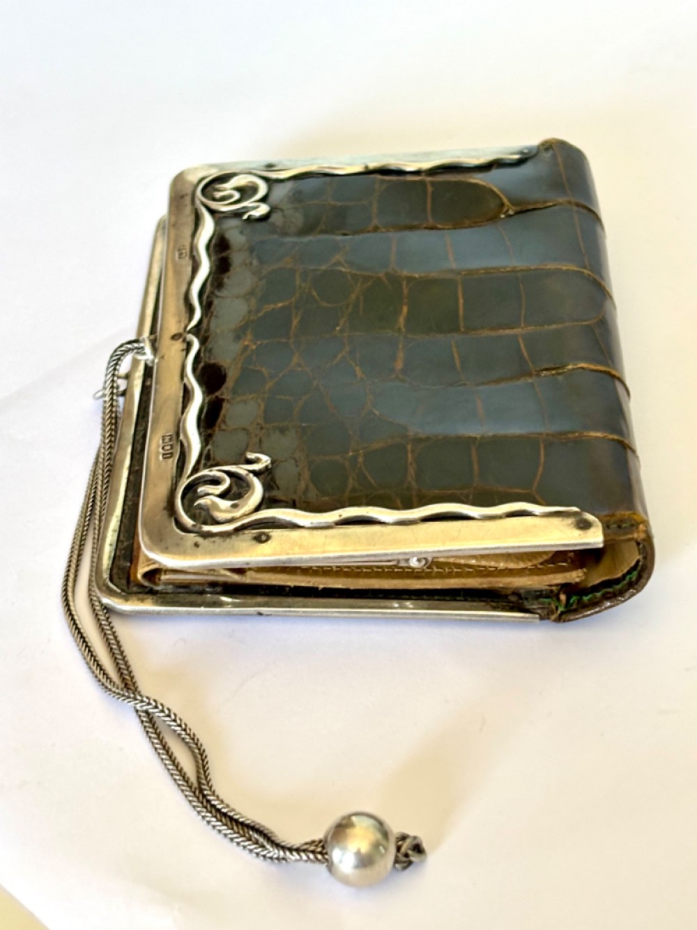 An Art Nouveau dark brown crocodile skin purse featuring hallmarked sterling silver whiplash corner mounts, a central silver cartouche with bow detailing, and a silver chain strap. View X