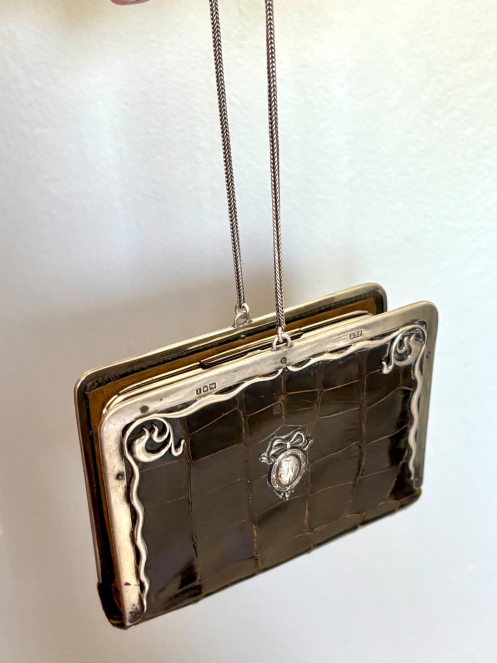 An Art Nouveau dark brown crocodile skin purse featuring hallmarked sterling silver whiplash corner mounts, a central silver cartouche with bow detailing, and a silver chain strap. View XV