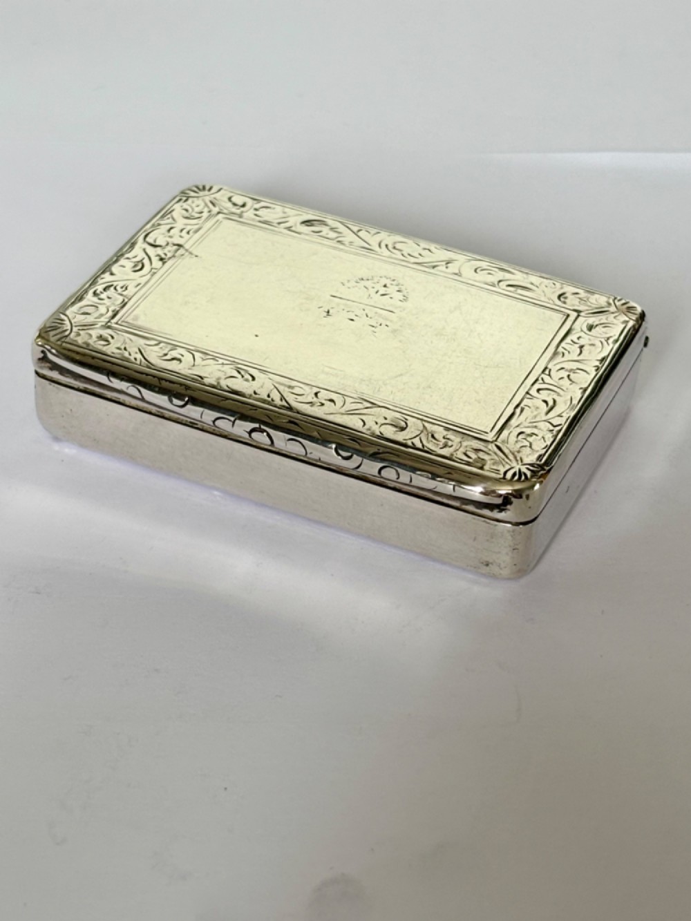 An early Victorian rectangular sterling silver gilt snuff box featuring engraved foliate scroll borders around a central crest on the hinged lid.
