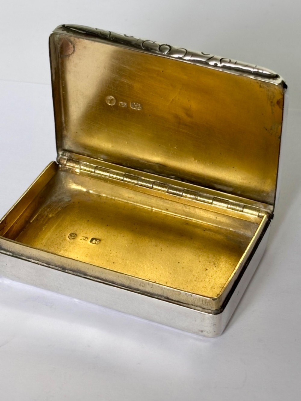 An early Victorian rectangular sterling silver gilt snuff box featuring engraved foliate scroll borders around a central crest on the hinged lid. View X