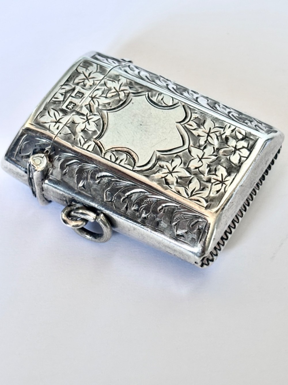 An engraved sterling silver vesta case featuring leaf motifs, a central vacant cartouche, a side suspension loop, and a base striker edge.