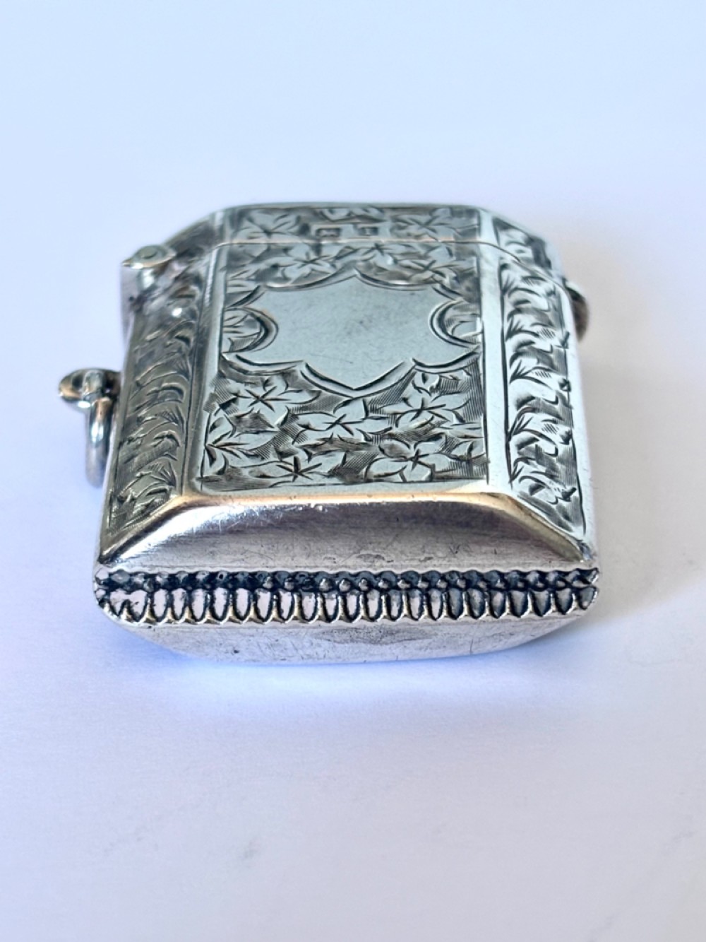 An engraved sterling silver vesta case featuring leaf motifs, a central vacant cartouche, a side suspension loop, and a base striker edge. View III