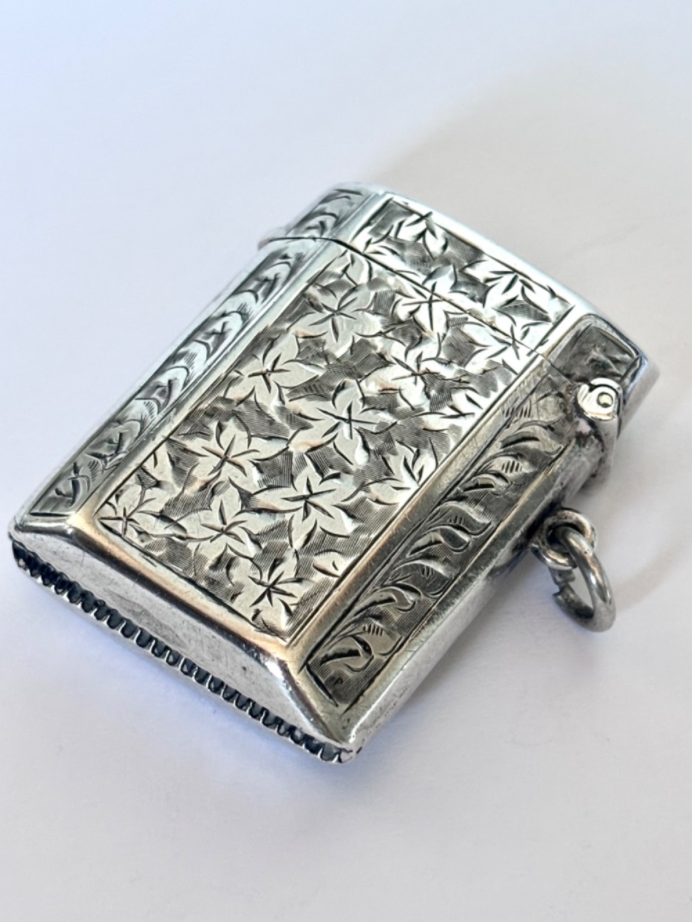 An engraved sterling silver vesta case featuring leaf motifs, a central vacant cartouche, a side suspension loop, and a base striker edge. View IV