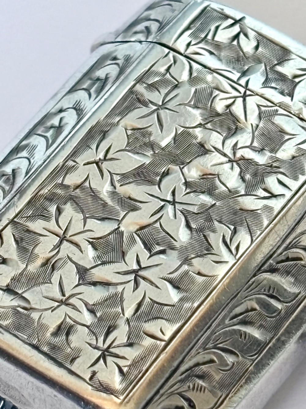 An engraved sterling silver vesta case featuring leaf motifs, a central vacant cartouche, a side suspension loop, and a base striker edge. View V