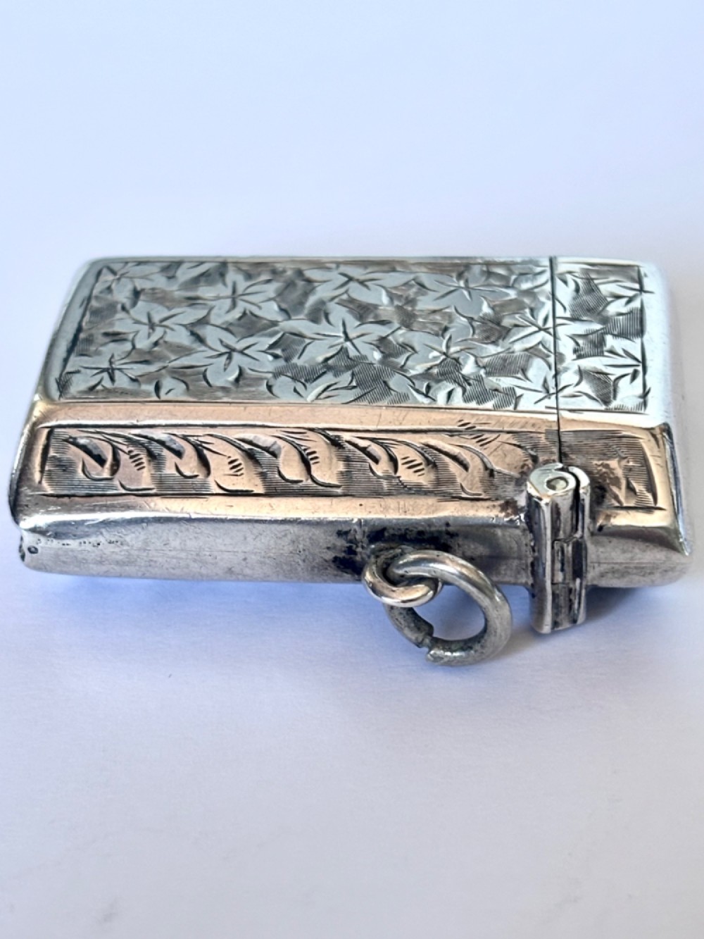 An engraved sterling silver vesta case featuring leaf motifs, a central vacant cartouche, a side suspension loop, and a base striker edge. View VI