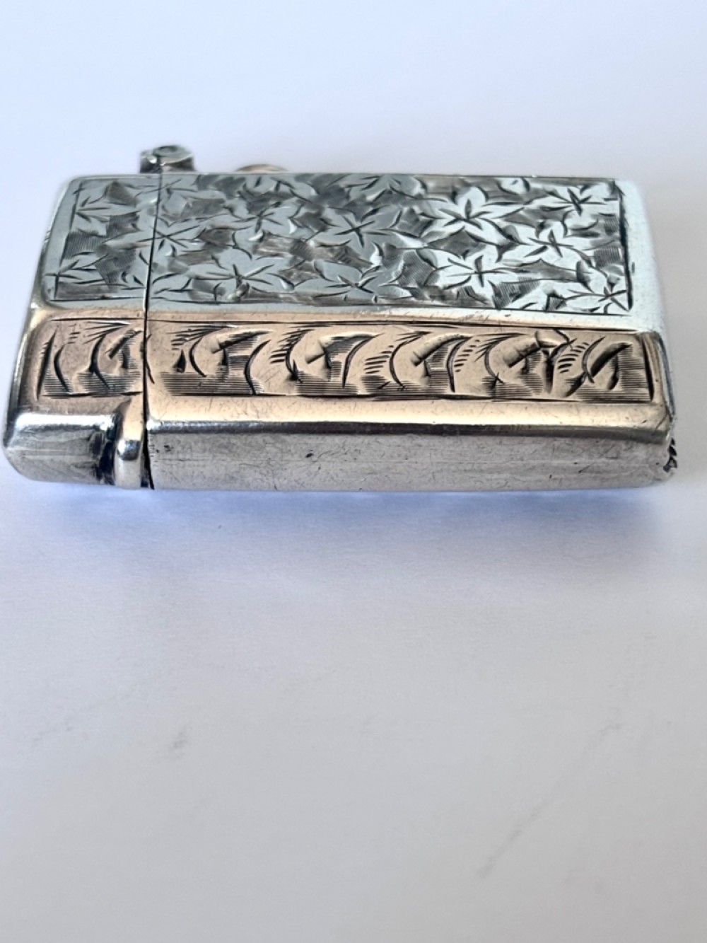 An engraved sterling silver vesta case featuring leaf motifs, a central vacant cartouche, a side suspension loop, and a base striker edge. View VII