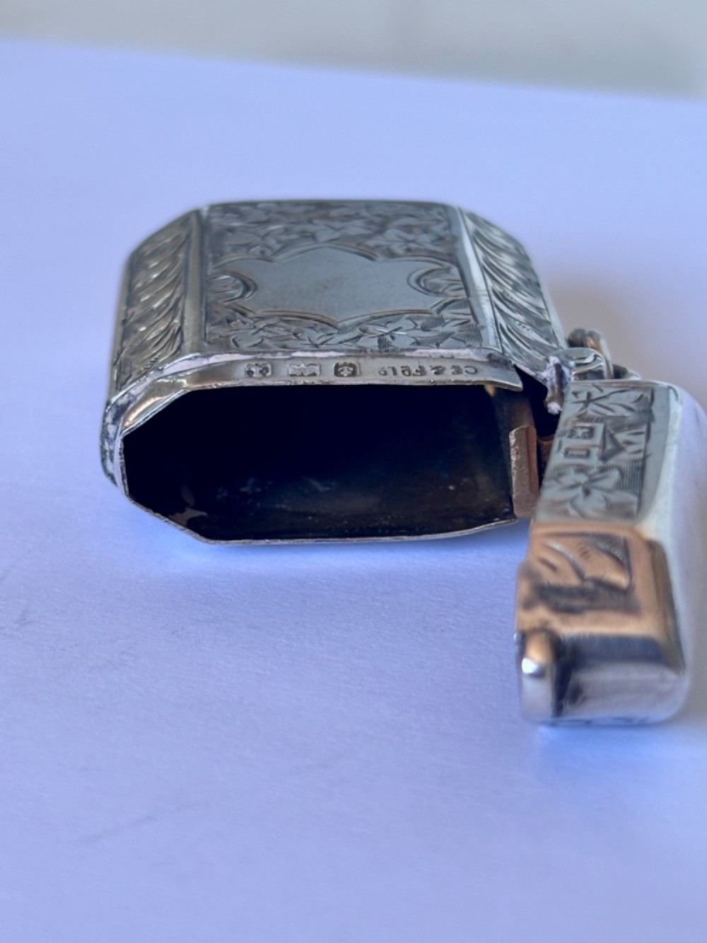 An engraved sterling silver vesta case featuring leaf motifs, a central vacant cartouche, a side suspension loop, and a base striker edge. View X