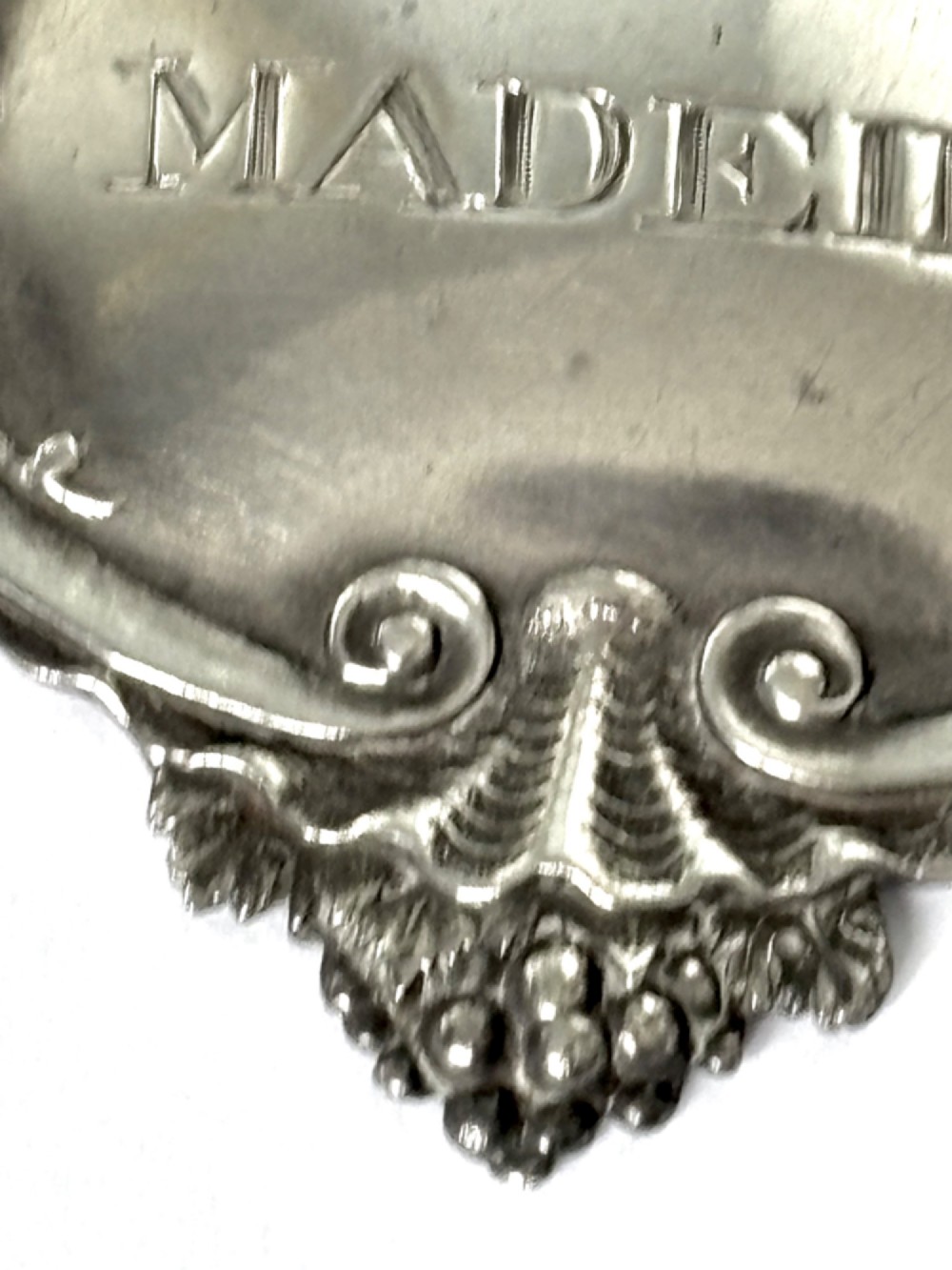 A George III sterling silver decanter label engraved with MADEIRA, decorated with a chased mask of Bacchus, grapevines, and an attached suspension chain. View V