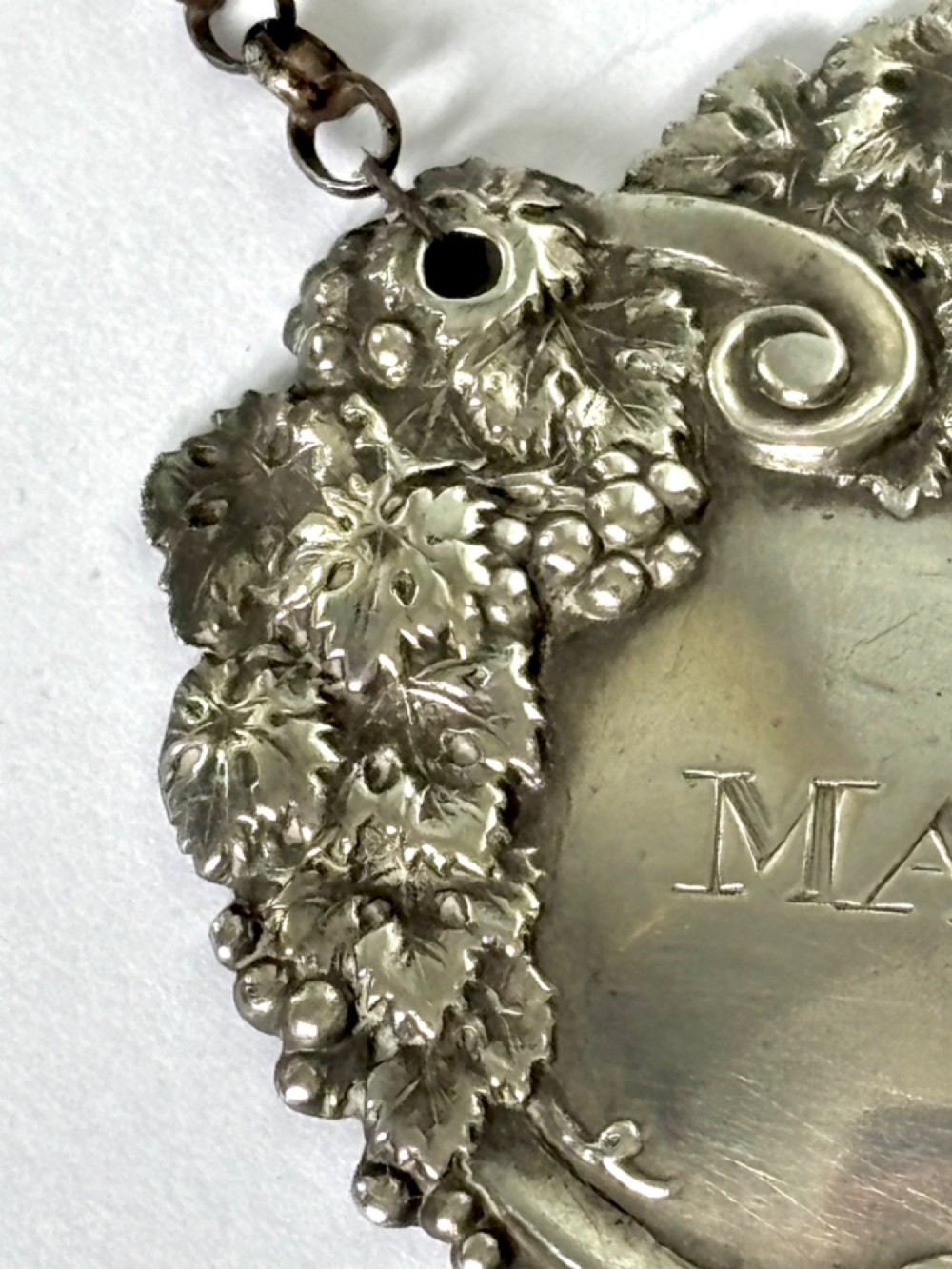 A George III sterling silver decanter label engraved with MADEIRA, decorated with a chased mask of Bacchus, grapevines, and an attached suspension chain. View VI