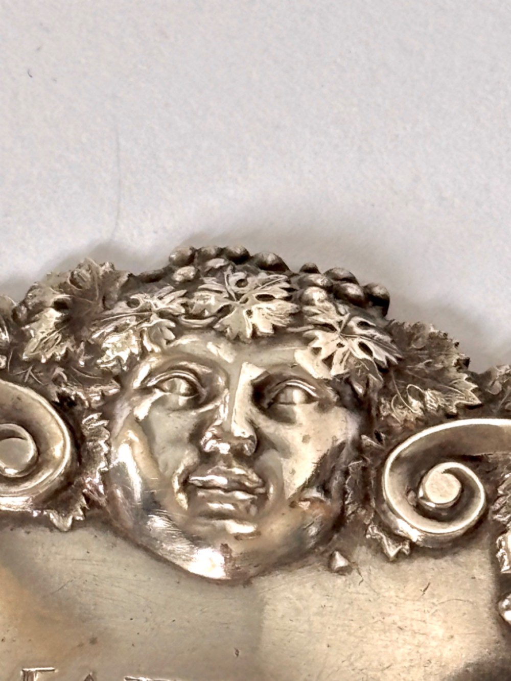 A George III sterling silver decanter label engraved with MADEIRA, decorated with a chased mask of Bacchus, grapevines, and an attached suspension chain. View VII