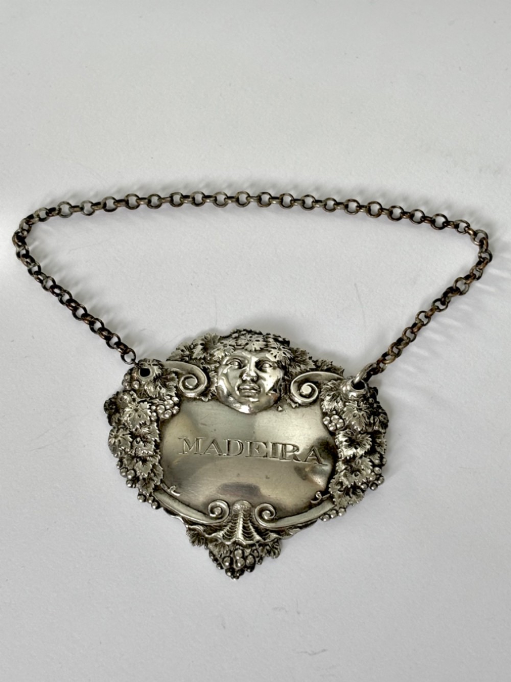 A George III sterling silver decanter label engraved with MADEIRA, decorated with a chased mask of Bacchus, grapevines, and an attached suspension chain. View VIII