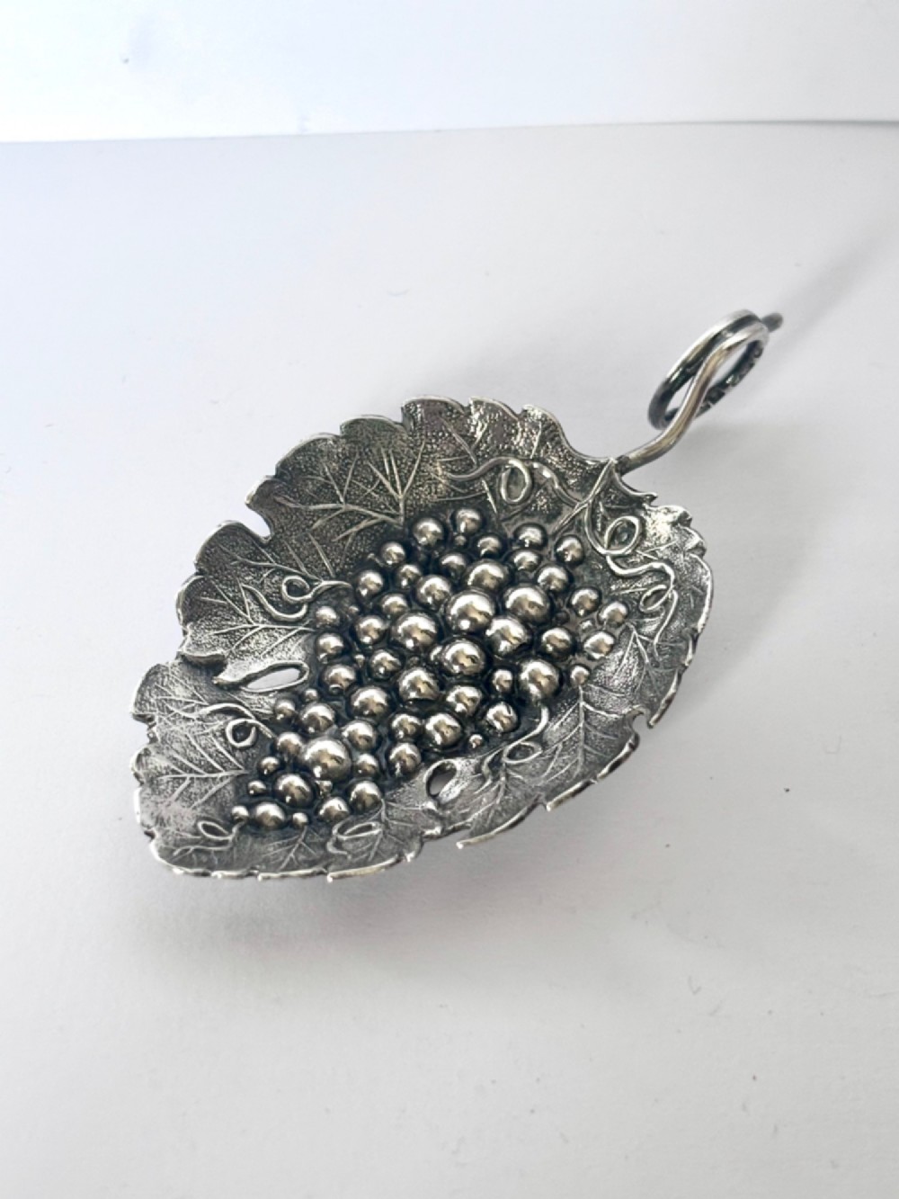 A George III sterling silver caddy spoon fashioned as a vine leaf with an embossed grape cluster and a wire loop handle.