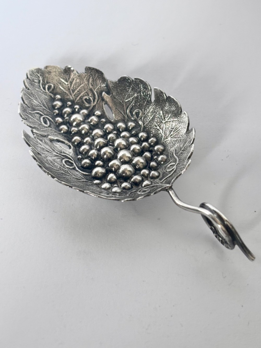 A George III sterling silver caddy spoon fashioned as a vine leaf with an embossed grape cluster and a wire loop handle. View II
