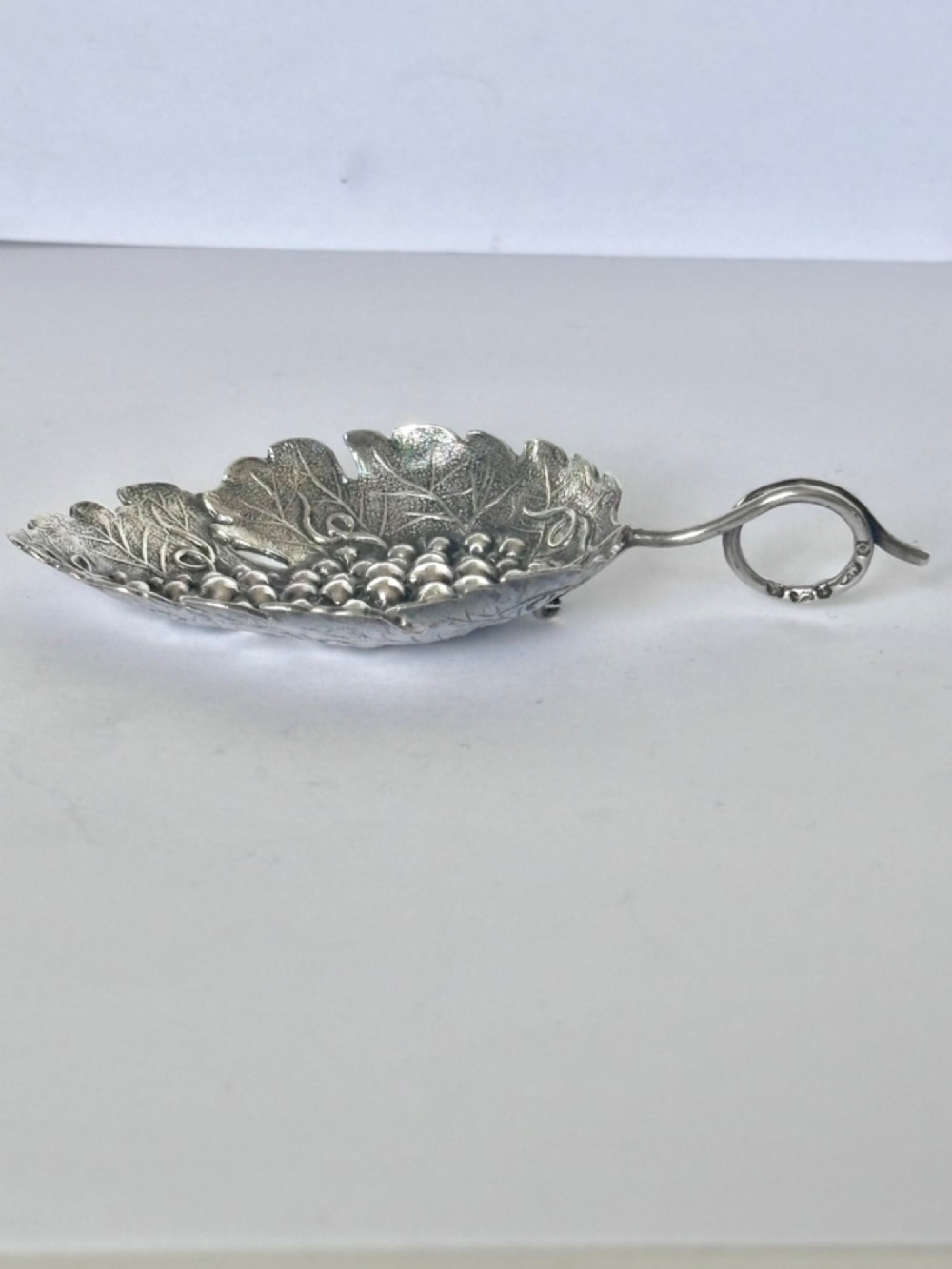 A George III sterling silver caddy spoon fashioned as a vine leaf with an embossed grape cluster and a wire loop handle. View X