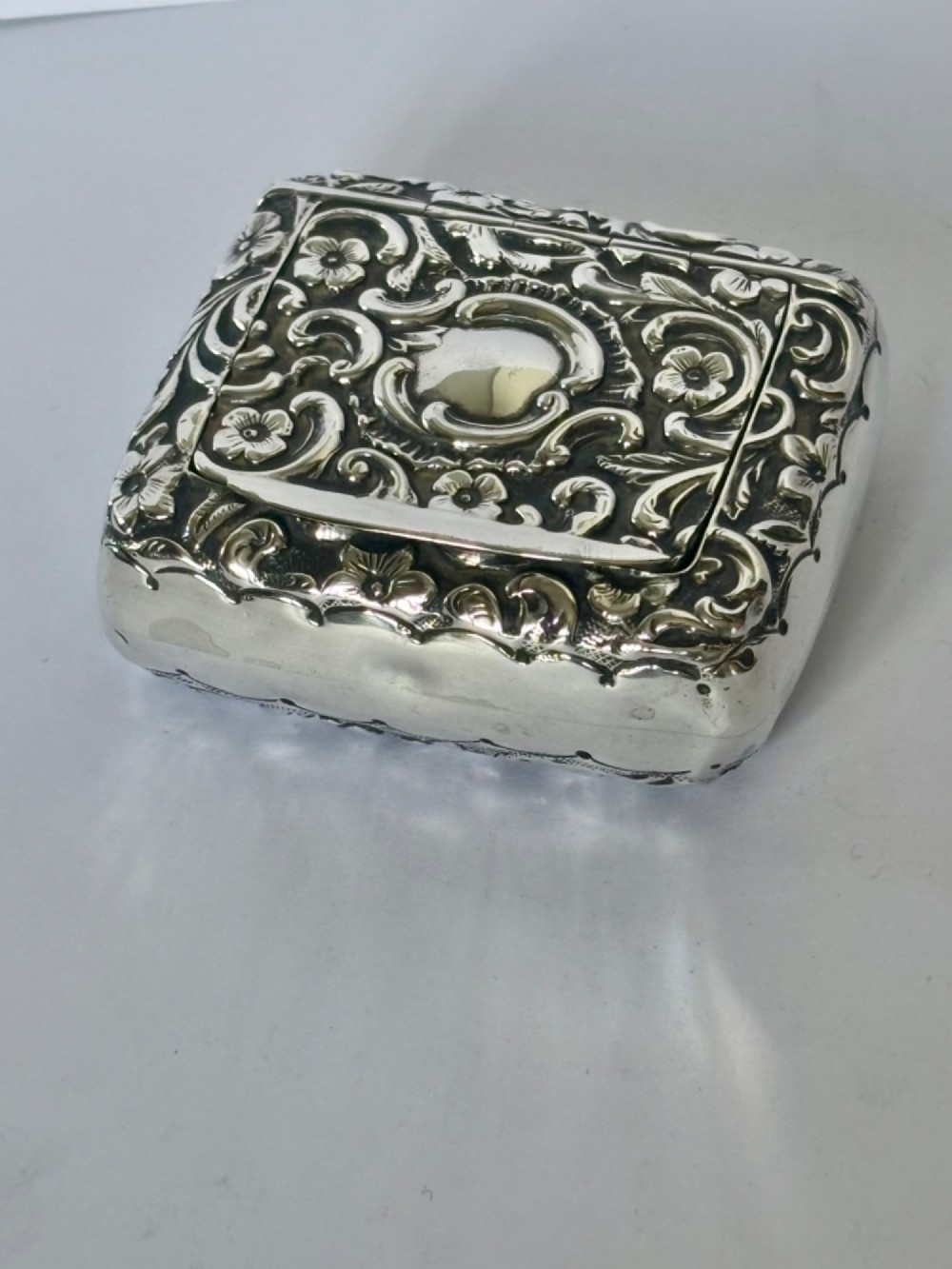 A Victorian rectangular sterling silver snuff box with heavily embossed floral and scroll decoration surrounding a plain central cartouche.