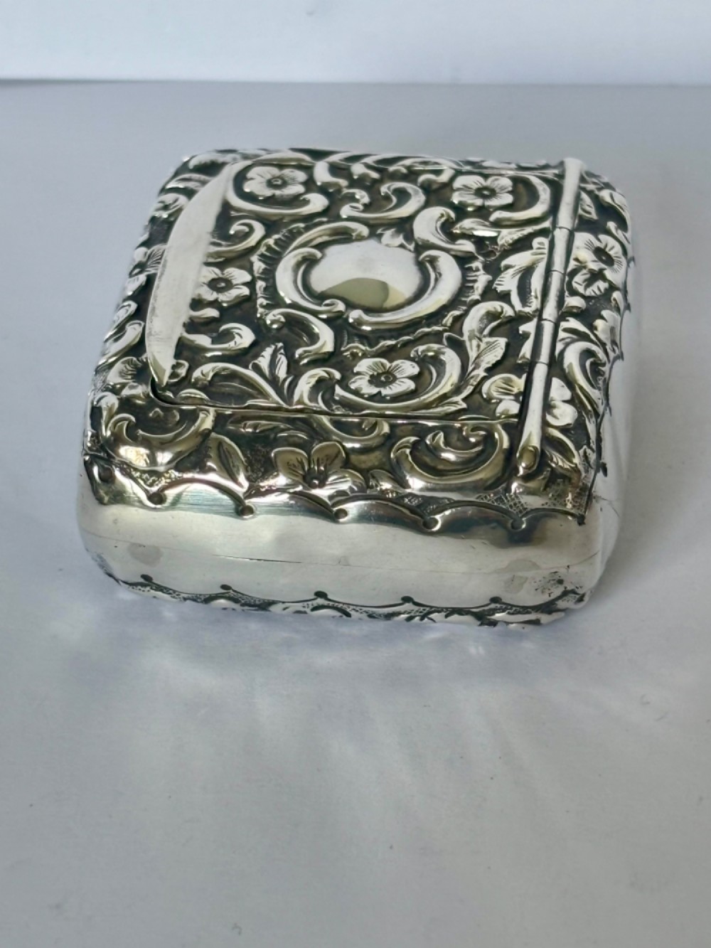 A Victorian rectangular sterling silver snuff box with heavily embossed floral and scroll decoration surrounding a plain central cartouche. View X