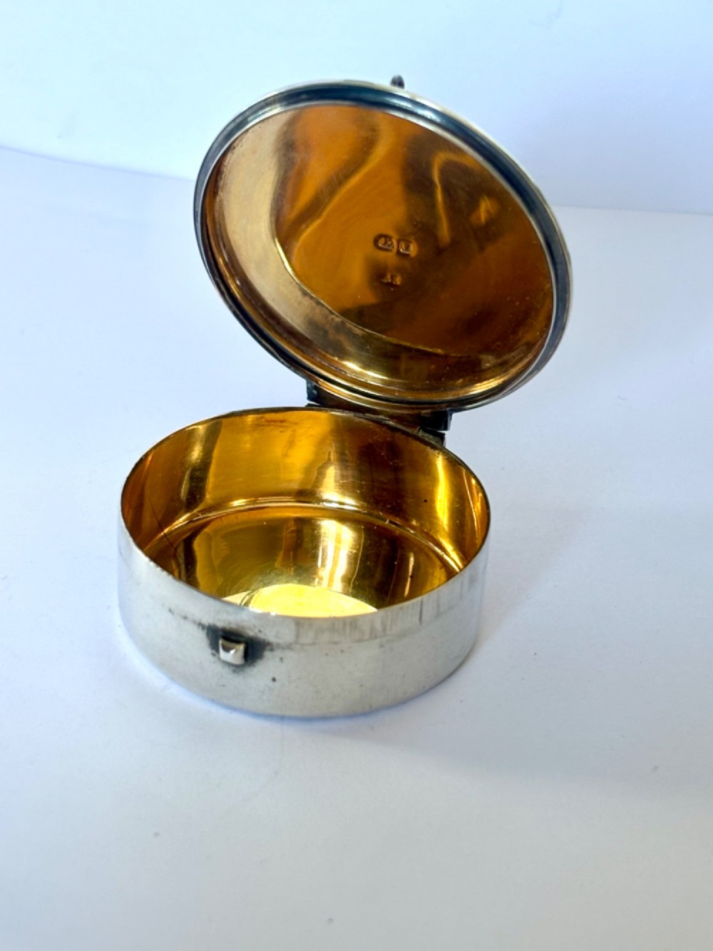A small round sterling silver pyx box shown open, featuring a silver exterior and a gold-gilt interior stamped with hallmarks.