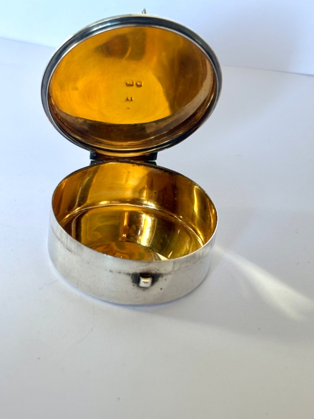 A small round sterling silver pyx box shown open, featuring a silver exterior and a gold-gilt interior stamped with hallmarks. View X