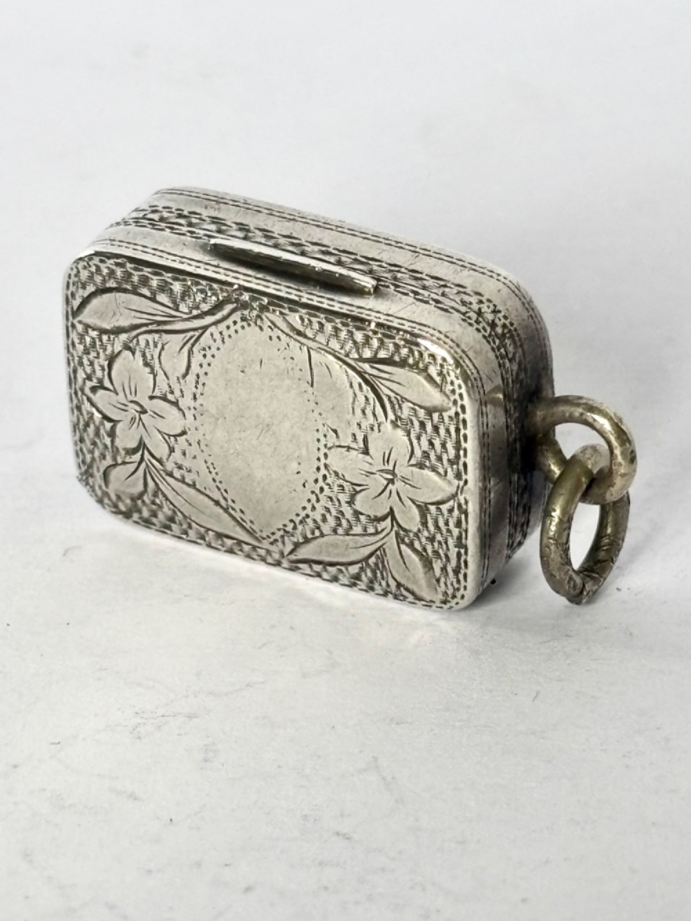 A small rectangular Georgian silver vinaigrette featuring intricate floral engraving, textured background patterns, and an attached suspension ring.