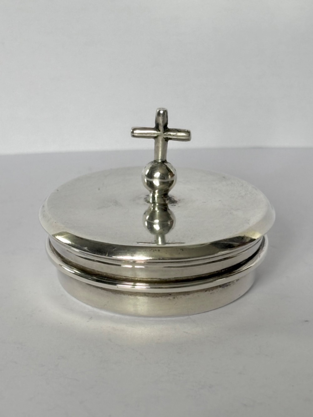 A circular sterling silver pyx box with a polished finish, featuring a lid topped with a cross and orb finial.