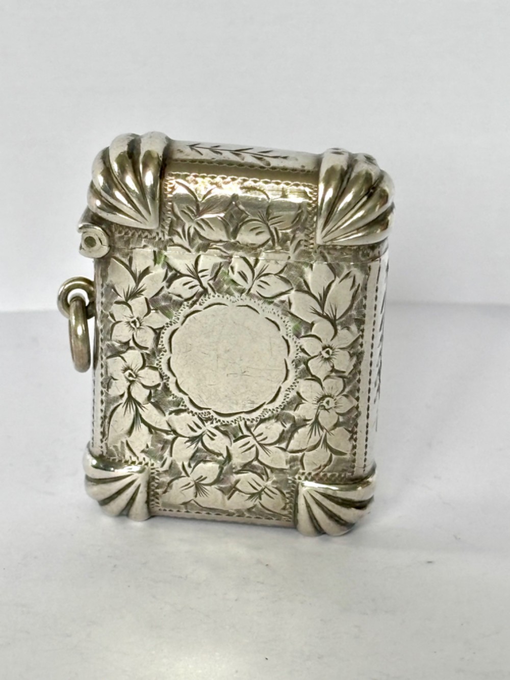 A Victorian sterling silver vesta case featuring hand-engraved floral leaf motifs, gadrooned corners, a vacant central cartouche, and a side suspension ring.