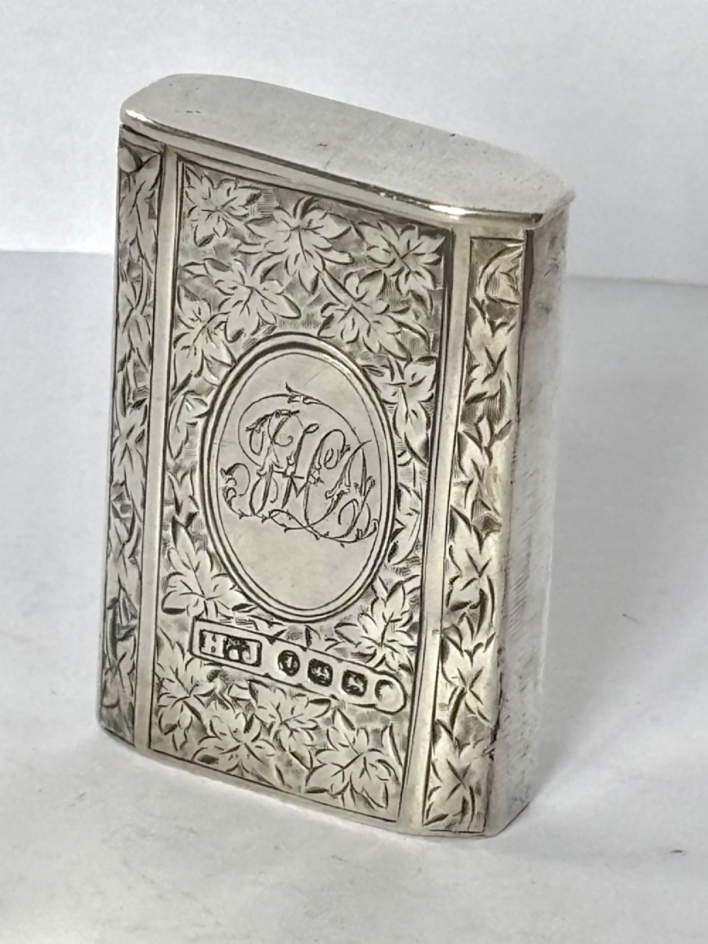 An upright rectangular Victorian sterling silver vesta case with hand-engraved leaf patterns, a central monogrammed oval cartouche, and prominent Birmingham hallmark stamps.