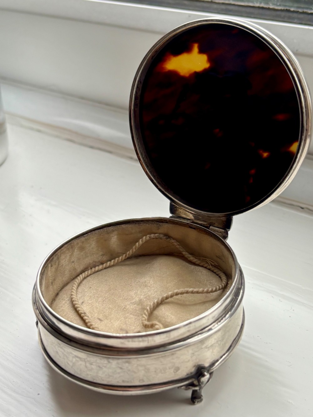 A circular silver dressing table box featuring a dark tortoiseshell lid with silver floral basket inlay resting on small shaped feet. View II