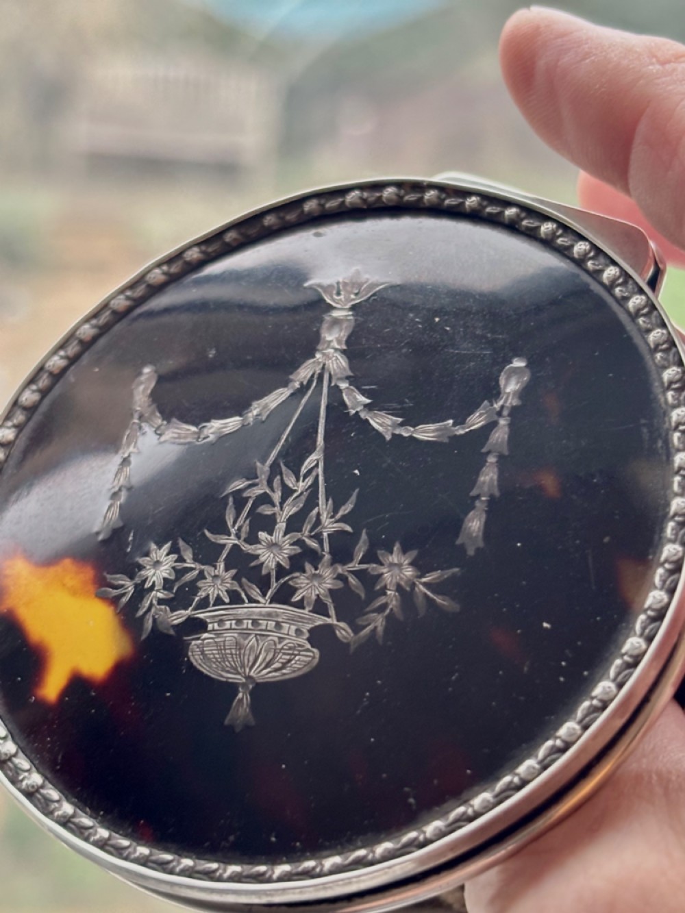 A circular silver dressing table box featuring a dark tortoiseshell lid with silver floral basket inlay resting on small shaped feet. View VII