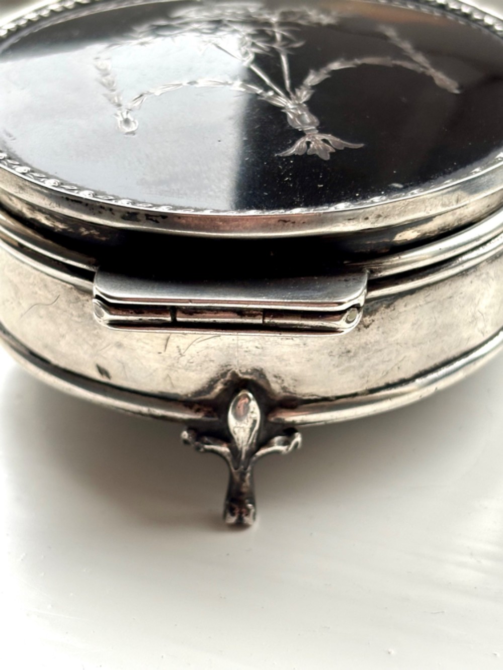 A circular silver dressing table box featuring a dark tortoiseshell lid with silver floral basket inlay resting on small shaped feet. View IX