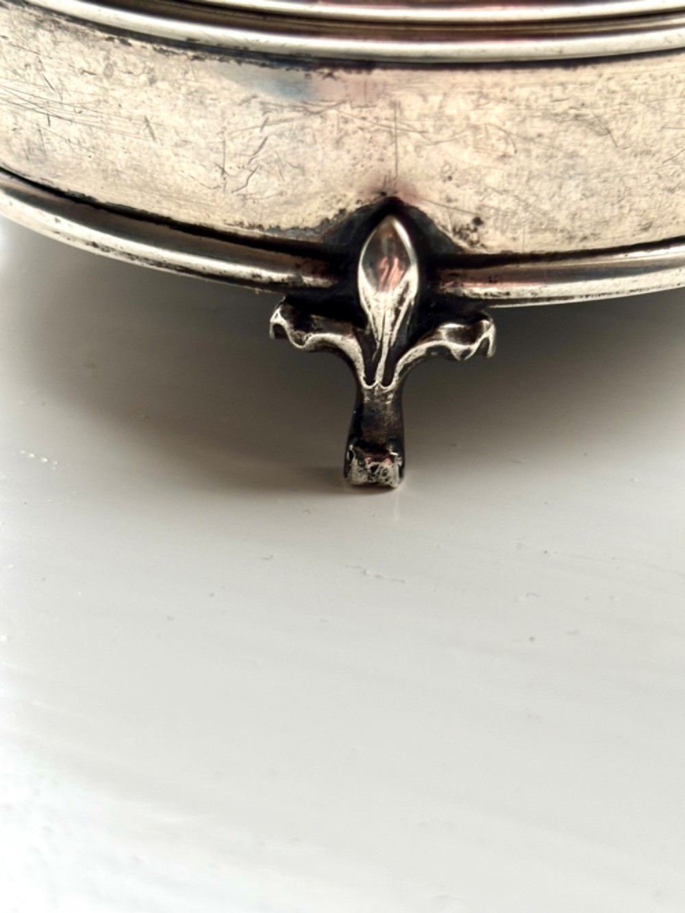A circular silver dressing table box featuring a dark tortoiseshell lid with silver floral basket inlay resting on small shaped feet. View X