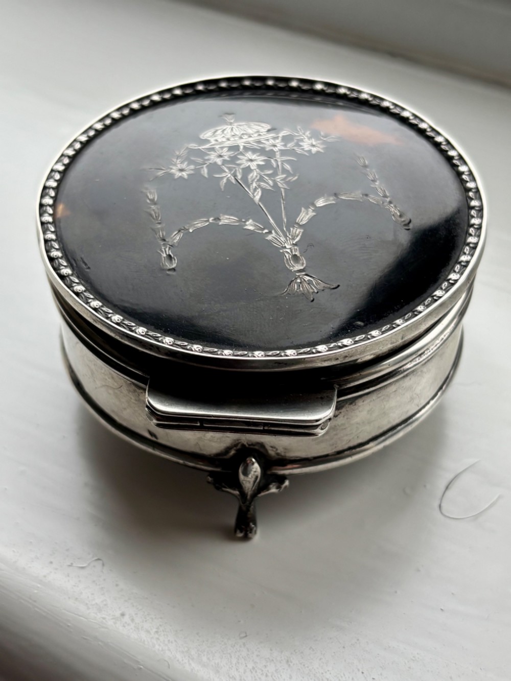 A circular silver dressing table box featuring a dark tortoiseshell lid with silver floral basket inlay resting on small shaped feet. View XI