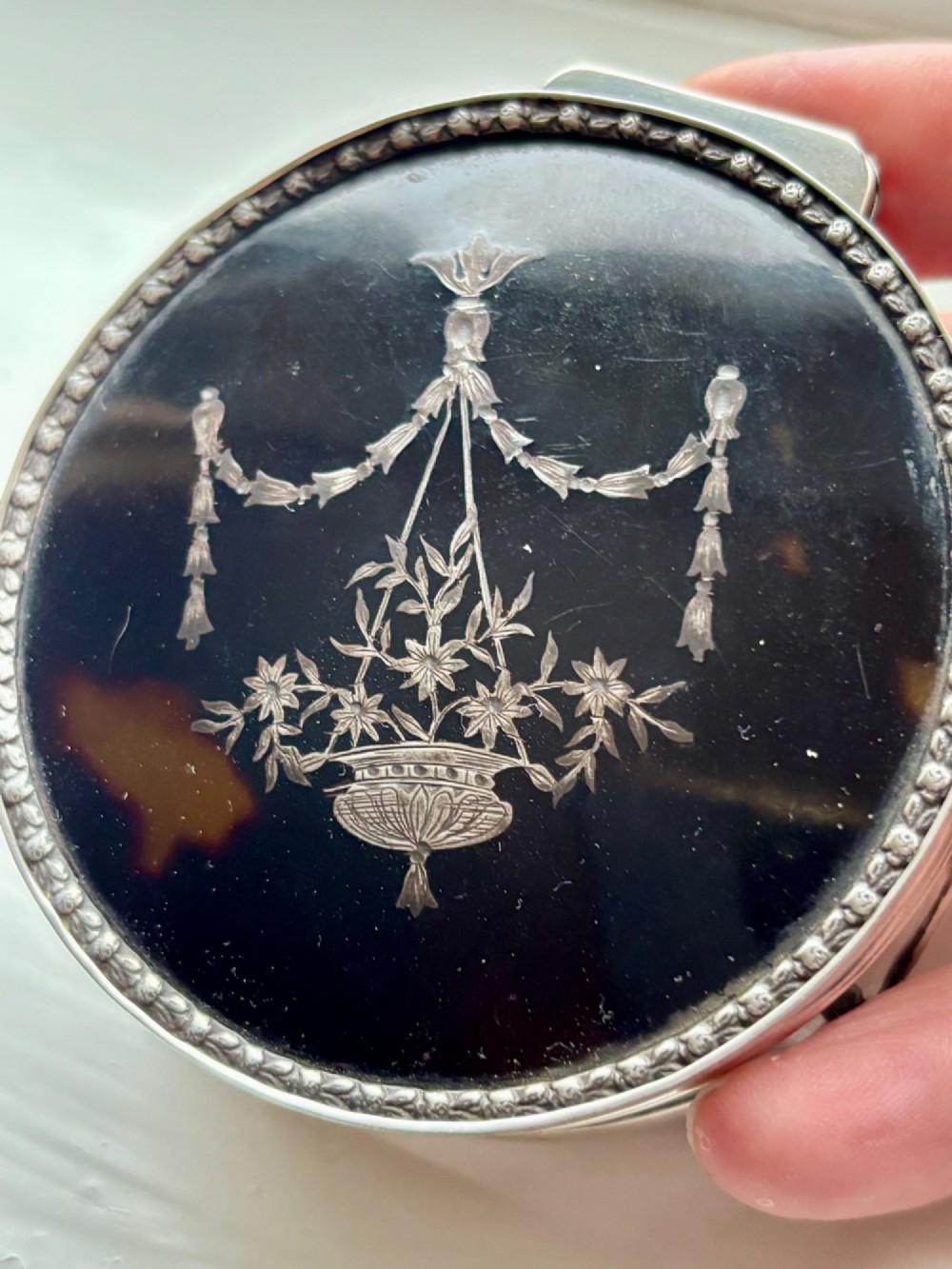A circular silver dressing table box featuring a dark tortoiseshell lid with silver floral basket inlay resting on small shaped feet. View XII