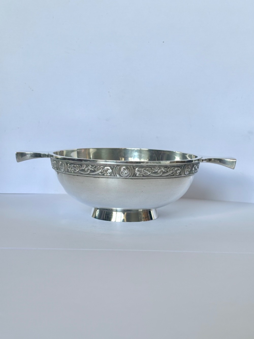 A shallow sterling silver quaich featuring two flattened lug handles, a circular pedestal base, and a band of embossed Celtic knotwork along the upper rim.