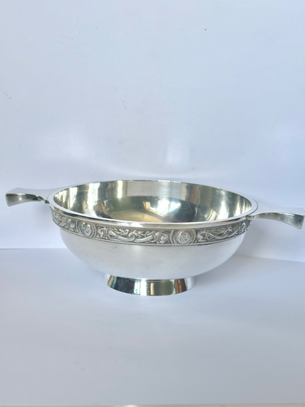 A shallow sterling silver quaich featuring two flattened lug handles, a circular pedestal base, and a band of embossed Celtic knotwork along the upper rim. View IV